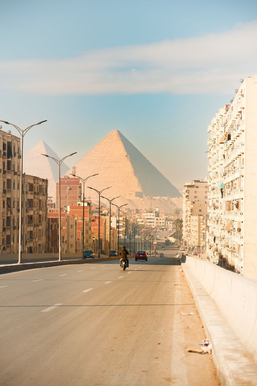Narrow street in Nazlet el-Samman with multi-story residential buildings, a pyramid behind them